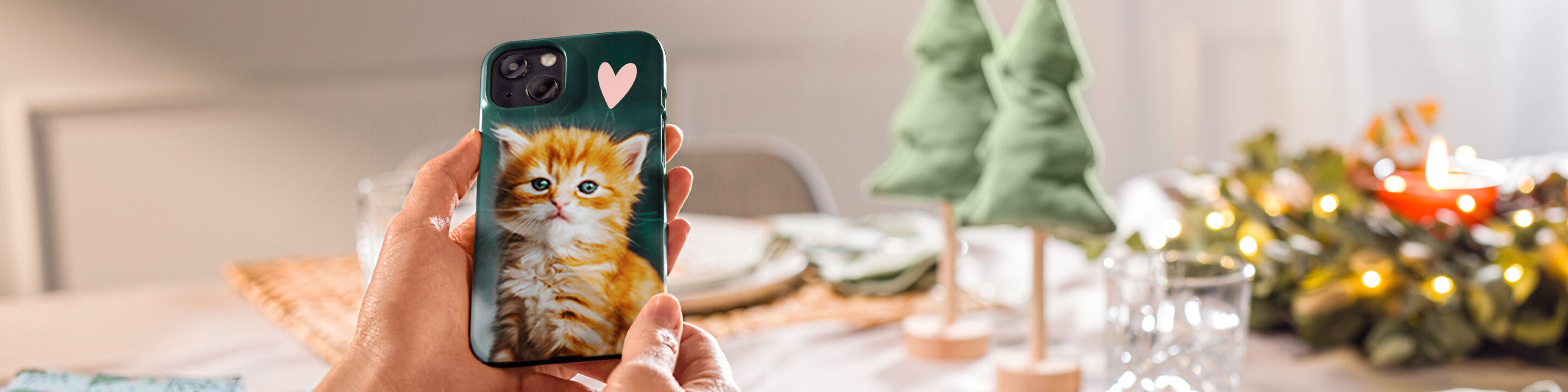 Little hands holding a smartphone displaying a cute kitten, in front of a festively decorated table scene.